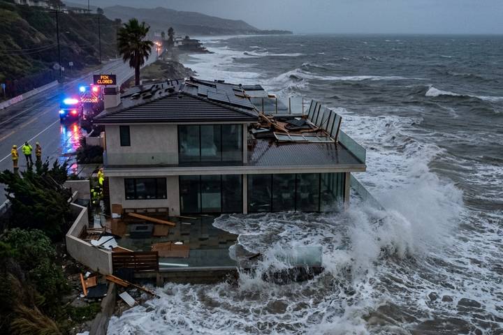 A coastal home with a damaged roof and scattered debris as storm waves hit the structure